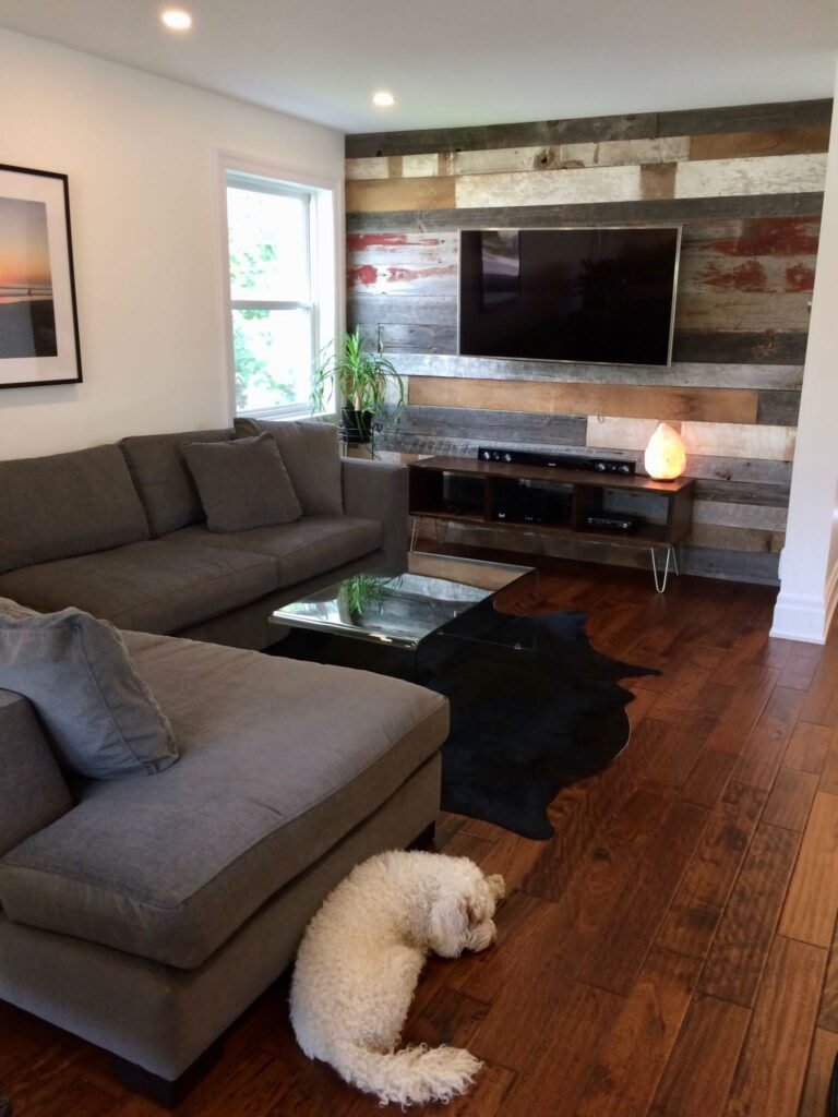Modern living room renovation with hardwood flooring, custom wood accent wall, wall-mounted TV, and stylish sectional sofa.